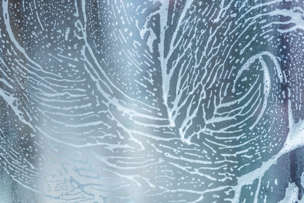 Traces de mousse de savon et gouttes d'eau sur une surface vitrée, formant des motifs ramifiés et des bulles dispersées. Fond bleu-gris flou.