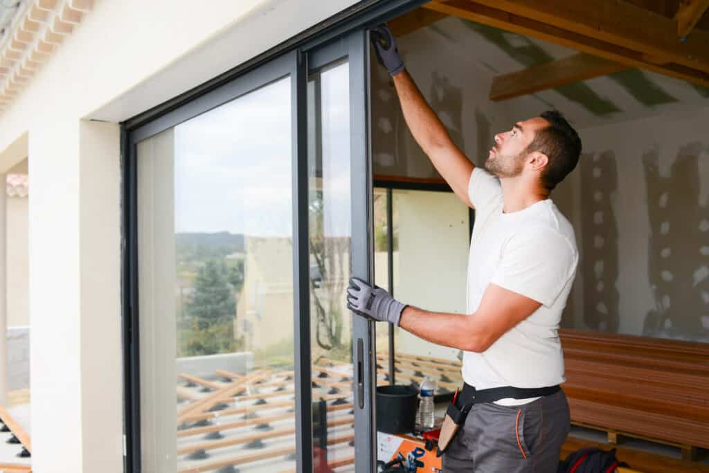 Poseur ajuste une grande porte coulissante vitrée noire. Intérieur en chantier avec murs bruts et poutres bois, extérieur vue sur construction de terrasse.